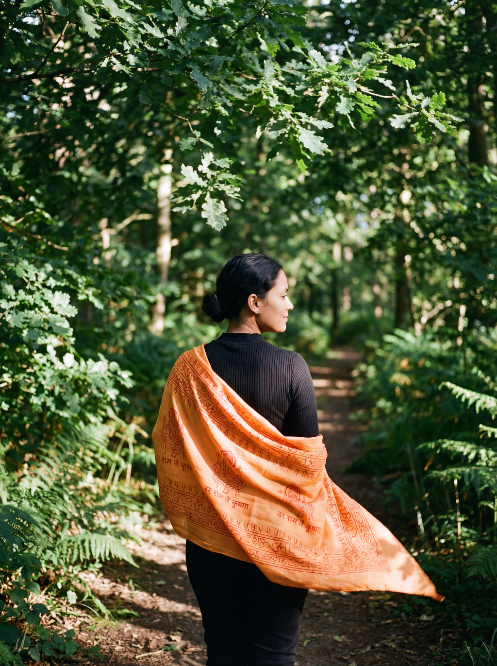 Woman with an orange print yoga shawl walking through a forest. The hand-dyed shawl is a Ganesha print pattern 