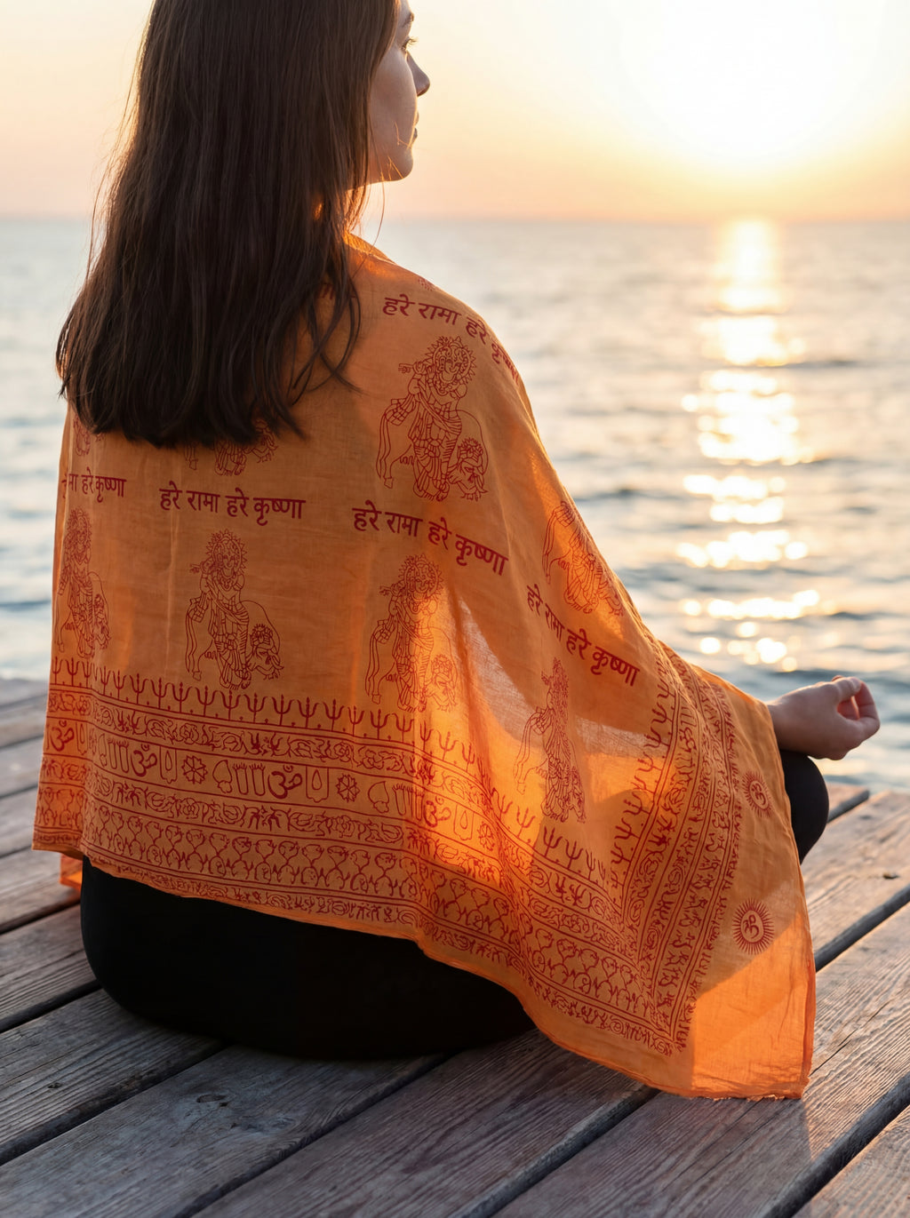 Person wearing an orange Krishna sanskrit patterned yoga shawl by the water at sunset