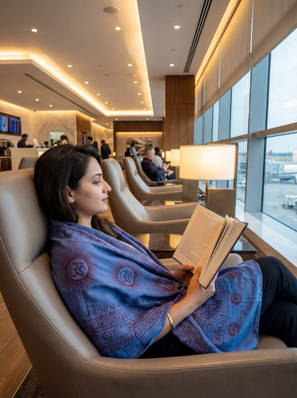 Woman wearing a blue Ram Nami yoga comfortable shawl reading a book in a modern airport lounge with large windows.